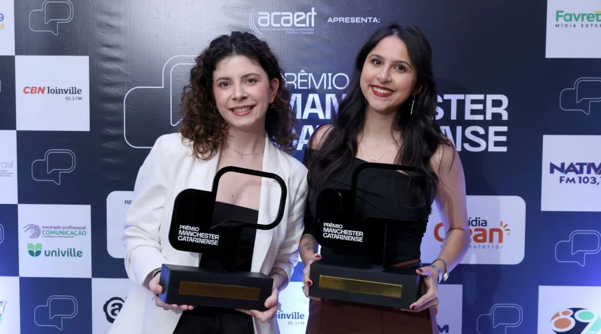 Bruna e Regielle sorrindo e segurando troféus do Prêmio Manchester Catarinense em frente a um painel com logotipos de patrocinadores e apoiadores do evento.