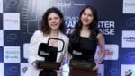 Bruna e Regielle sorrindo e segurando troféus do Prêmio Manchester Catarinense em frente a um painel com logotipos de patrocinadores e apoiadores do evento.