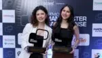 Bruna e Regielle sorrindo e segurando troféus do Prêmio Manchester Catarinense em frente a um painel com logotipos de patrocinadores e apoiadores do evento.