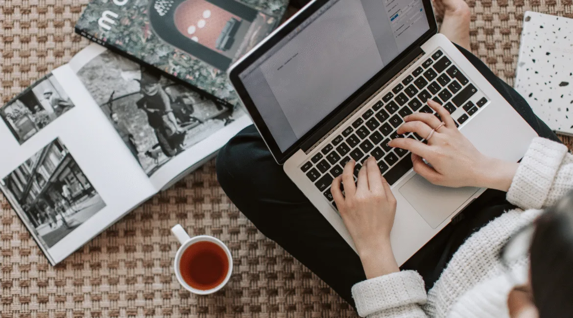 Pessoa digitando em um notebook, com livros e uma xícara de chá ao lado, representando produção de conteúdo digital para empresa B2B.
