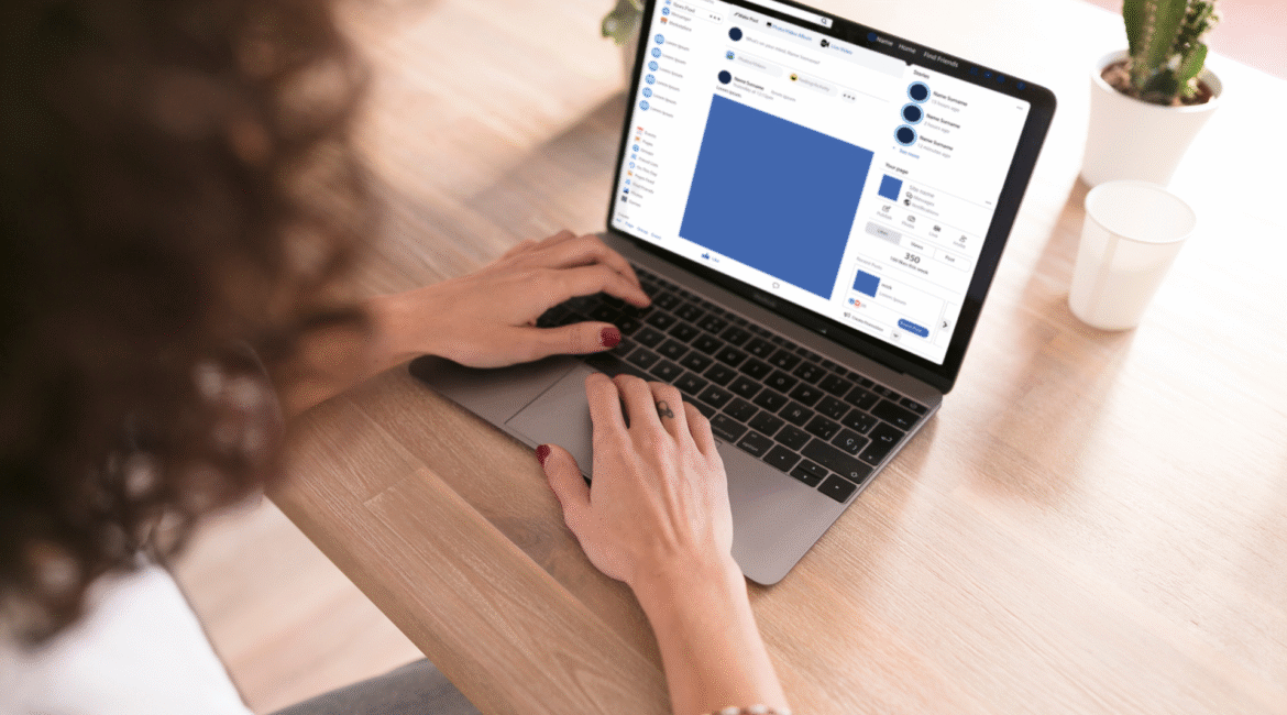 Mulher usando um notebook em uma mesa de madeira, navegando no LinkedIn, com mãos sobre o teclado e uma planta pequena ao lado.