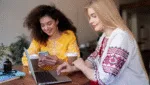 Duas profissionais interagindo com notebook e celular em um trabalhando em um café, representando o conceito de social selling e construção de conexões no ambiente digital.