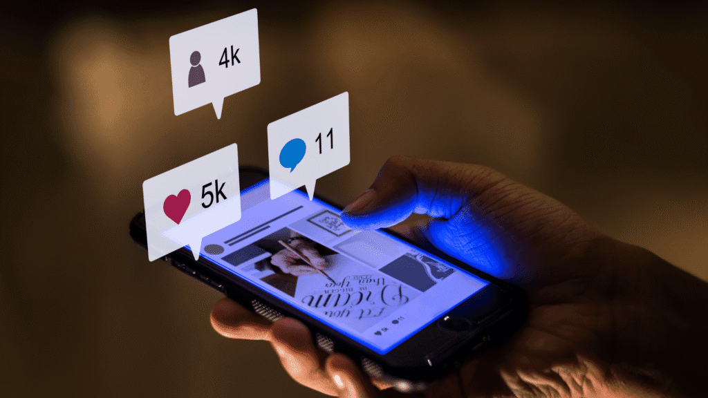 Mão segurando um smartphone com uma publicação aberta, enquanto ícones de curtidas, comentários e novos seguidores aparecem acima da tela, simbolizando crescimento e engajamento na presença orgânica nas redes sociais.