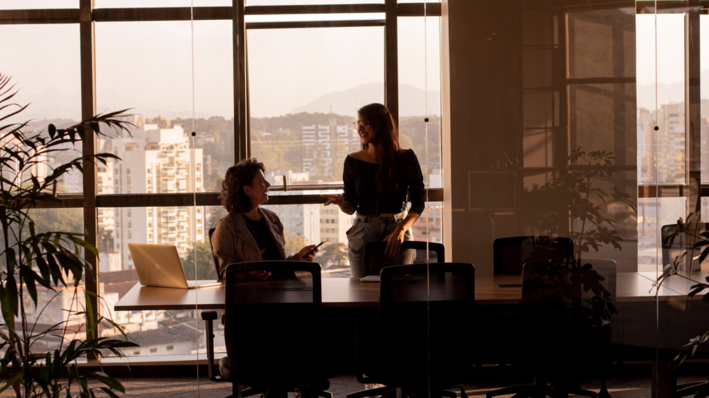 Escritório físico moderno com grandes janelas, duas mulheres conversando ao redor de uma mesa de trabalho, laptops abertos e vista urbana ao fundo, transmitindo profissionalismo e estratégia.