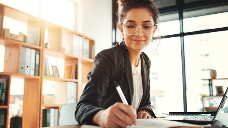 Advogada escrevendo em um escritório moderno enquanto produz conteúdo especializado, representando estratégias de marketing jurídico voltadas ao fortalecimento da autoridade, da credibilidade e do relacionamento com potenciais clientes.
